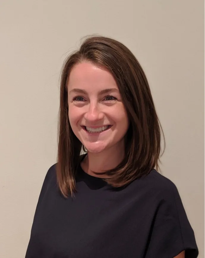 Woman with brown hair smiling, wearing dark blue shirt against neutral background