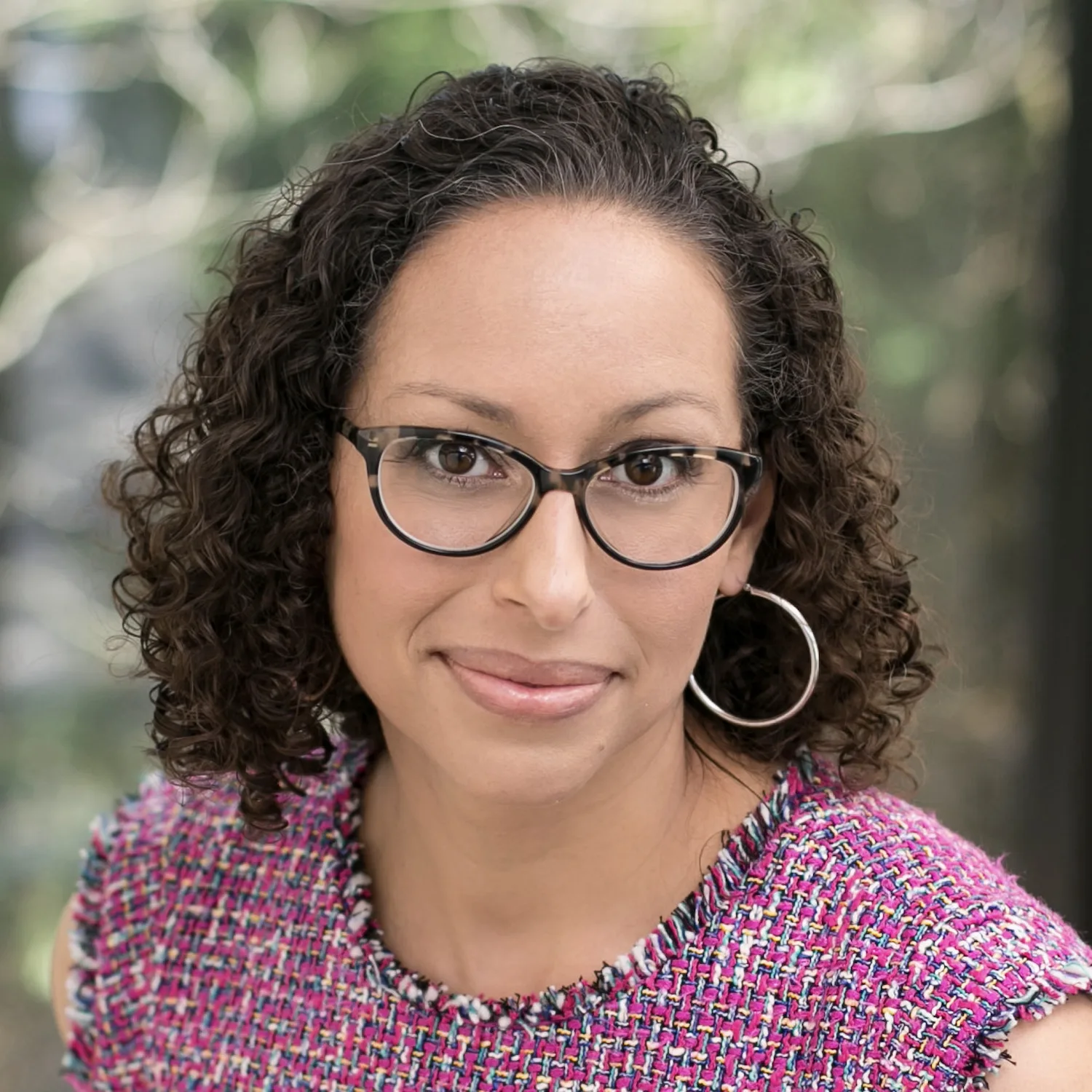 Woman with curly hair, glasses, and hoop earrings wearing colorful textured top