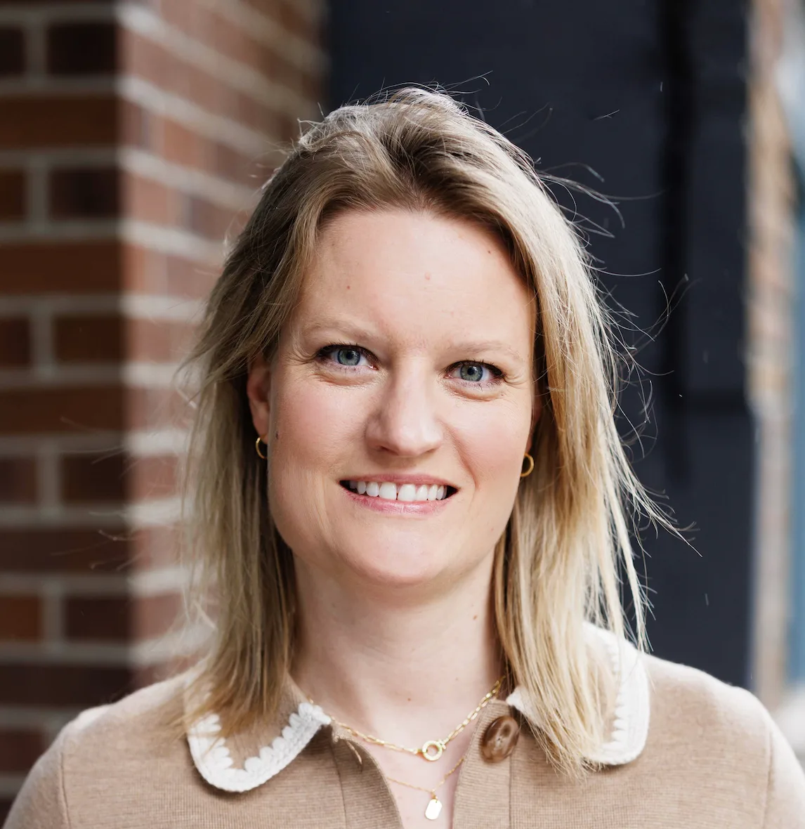 Woman with blonde hair and blue eyes smiling in front of brick wall