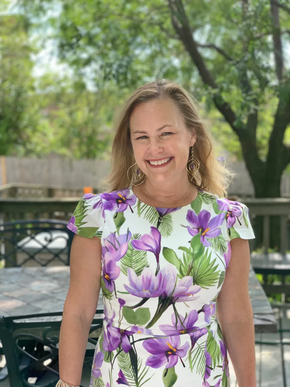 Woman in purple floral dress smiling outdoors with large hoop earrings