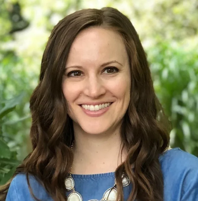 Woman with brown wavy hair wearing blue top and white stone necklace smiling outdoors
