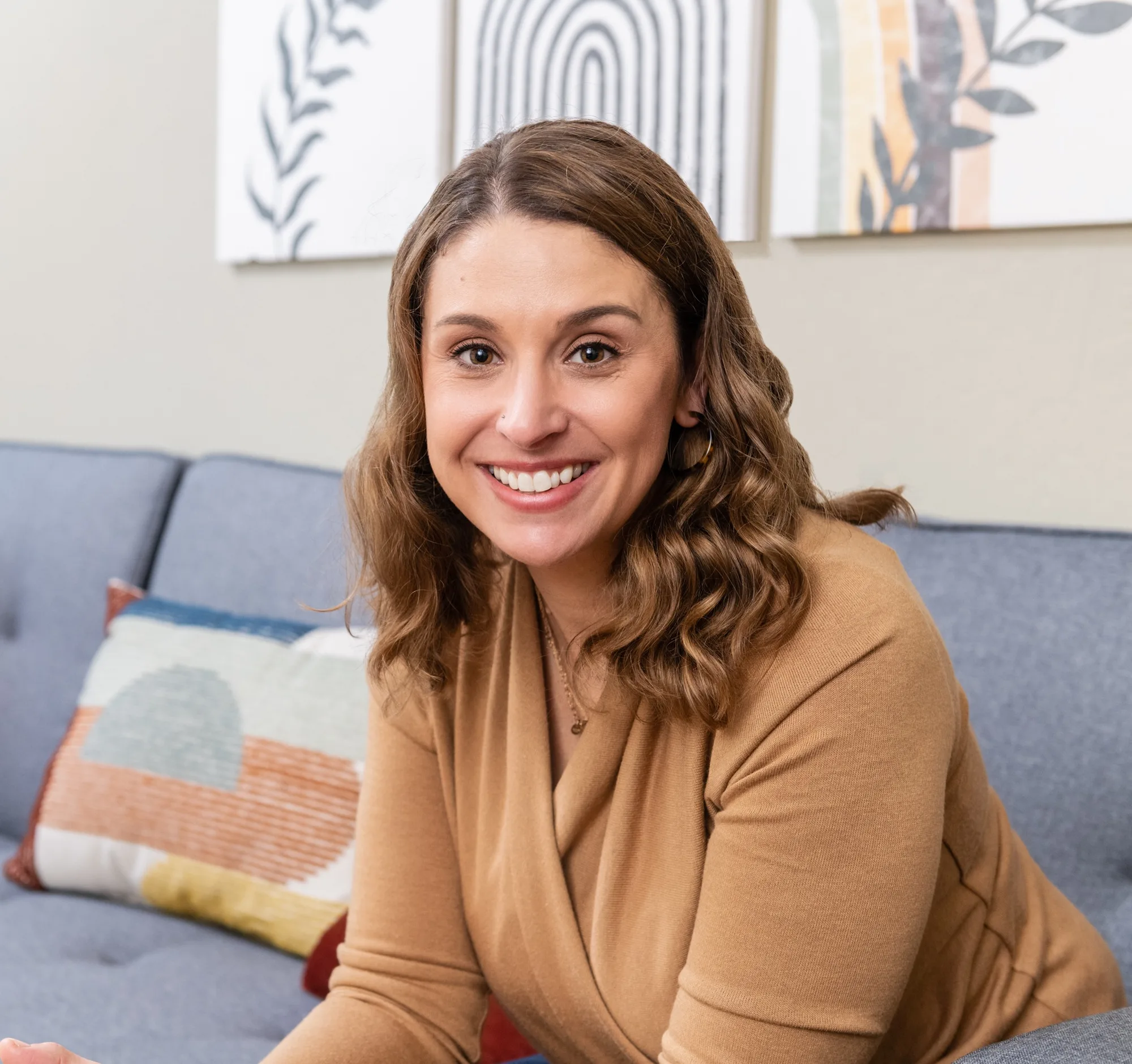 Woman with wavy brown hair in tan sweater smiling on gray couch