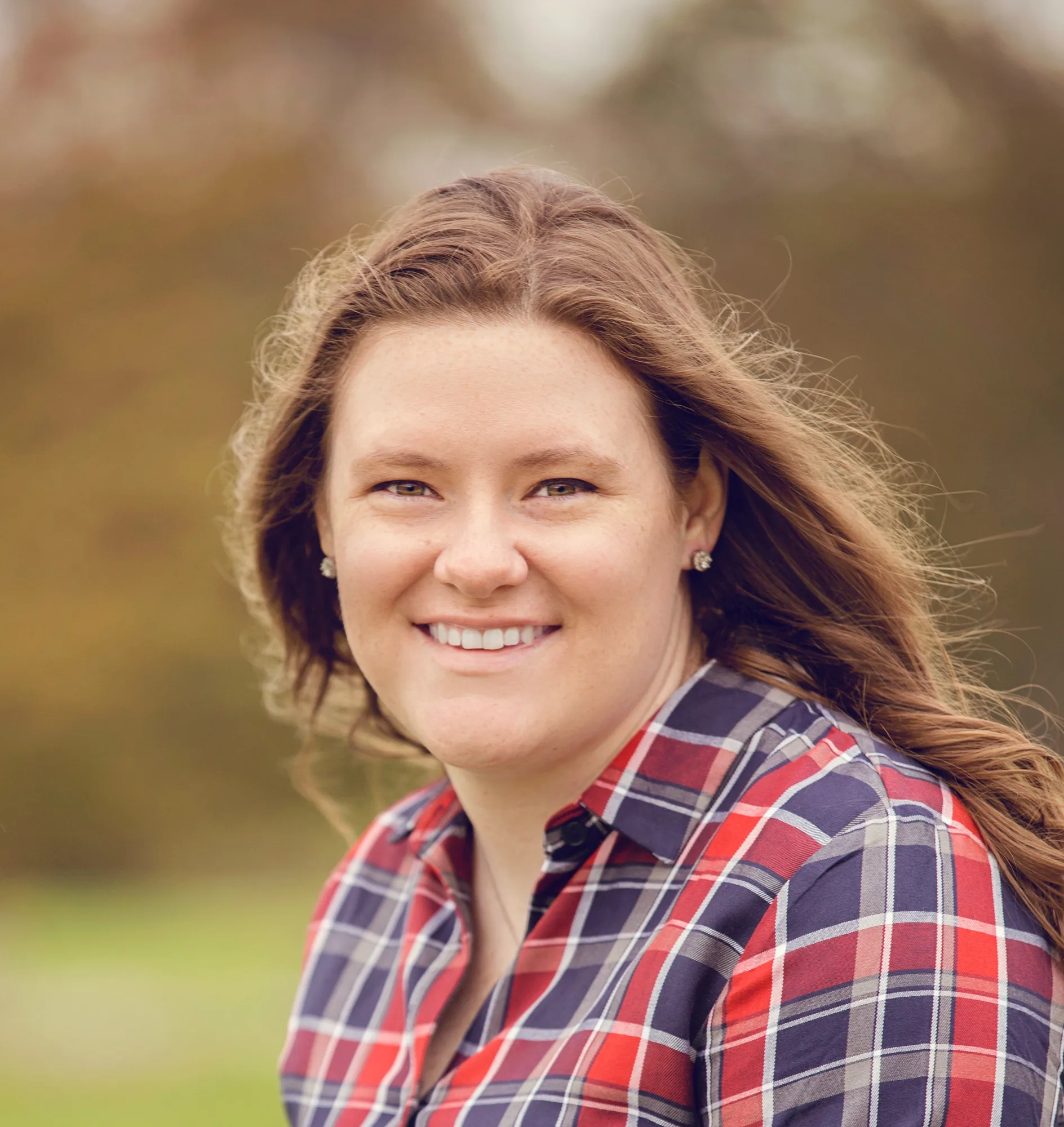 Woman with long brown hair in red and navy plaid shirt smiling outdoors