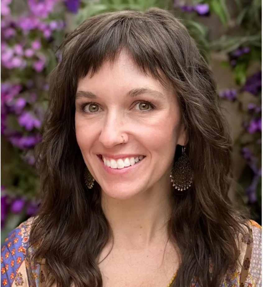 Woman with wavy brown hair and decorative earrings smiling in front of purple flowers