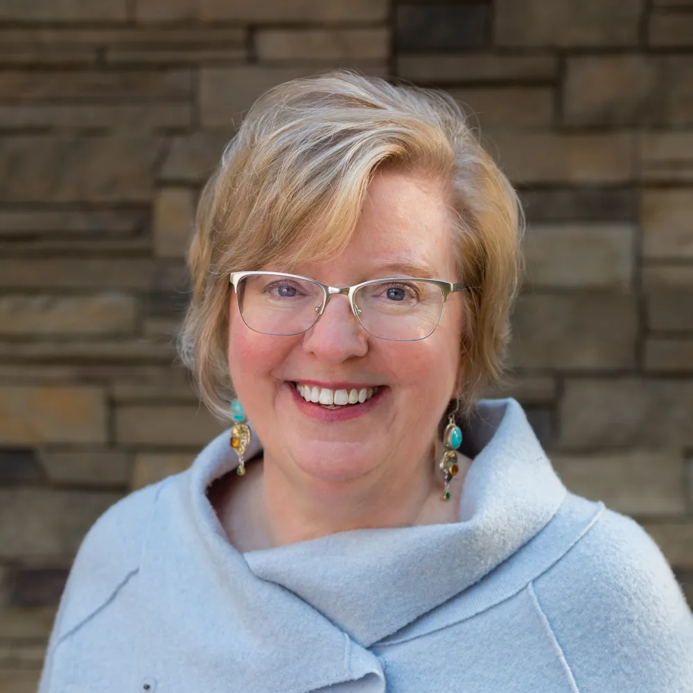 Woman with blonde hair and glasses wearing light blue top with decorative earrings