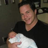 Man in black v-neck shirt smiling while holding a newborn baby wrapped in white