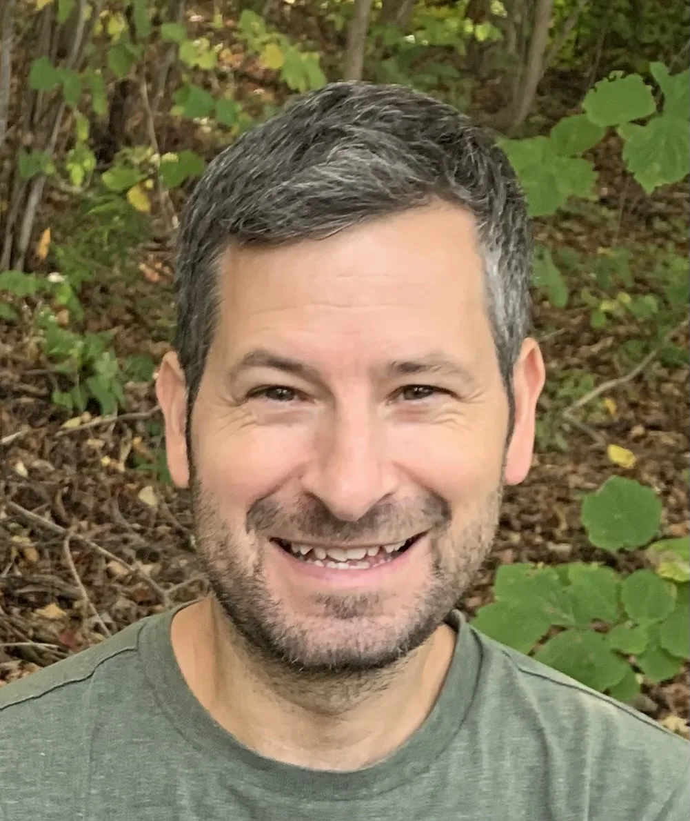 Man with gray hair and beard smiling outdoors in wooded area