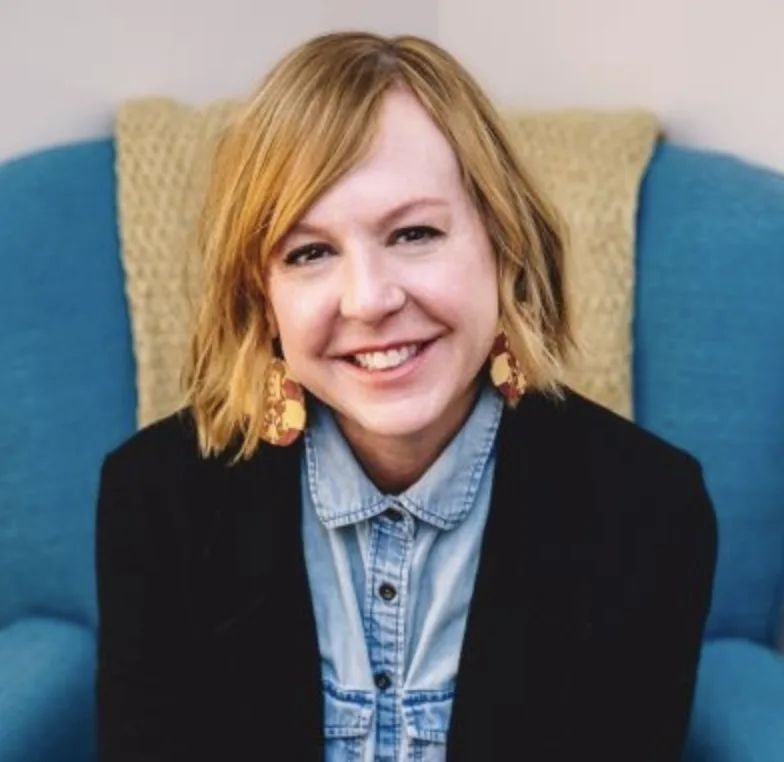 Woman with blonde hair wearing gold earrings, denim shirt, and black vest, smiling at camera