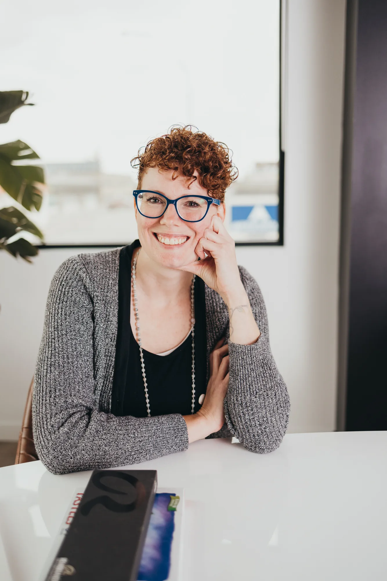 Smiling woman with curly hair and blue glasses at desk in bright office
