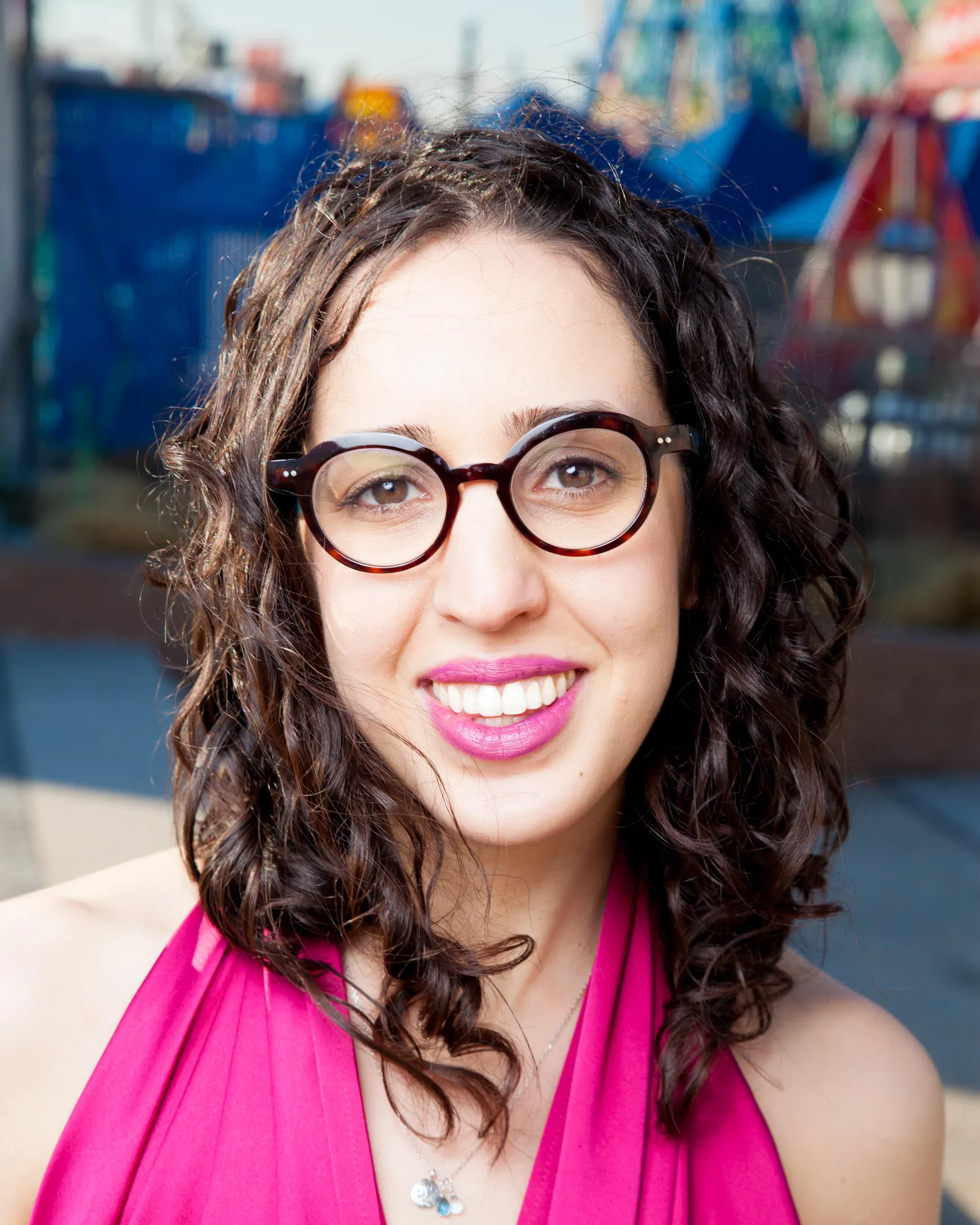 Woman with curly dark hair, round glasses, and bright pink lipstick smiling at camera