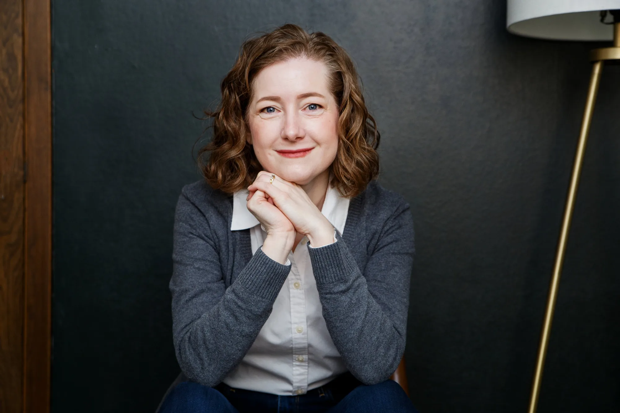 Woman with wavy brown hair wearing gray cardigan and white shirt, hands folded under chin