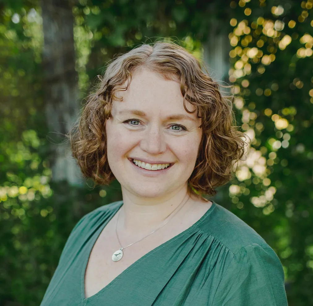 Woman with curly auburn hair wearing green dress and heart necklace smiling outdoors