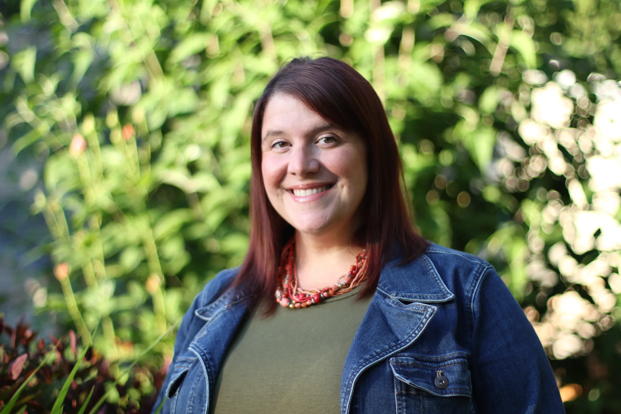Woman in denim jacket with red beaded necklace smiling outdoors