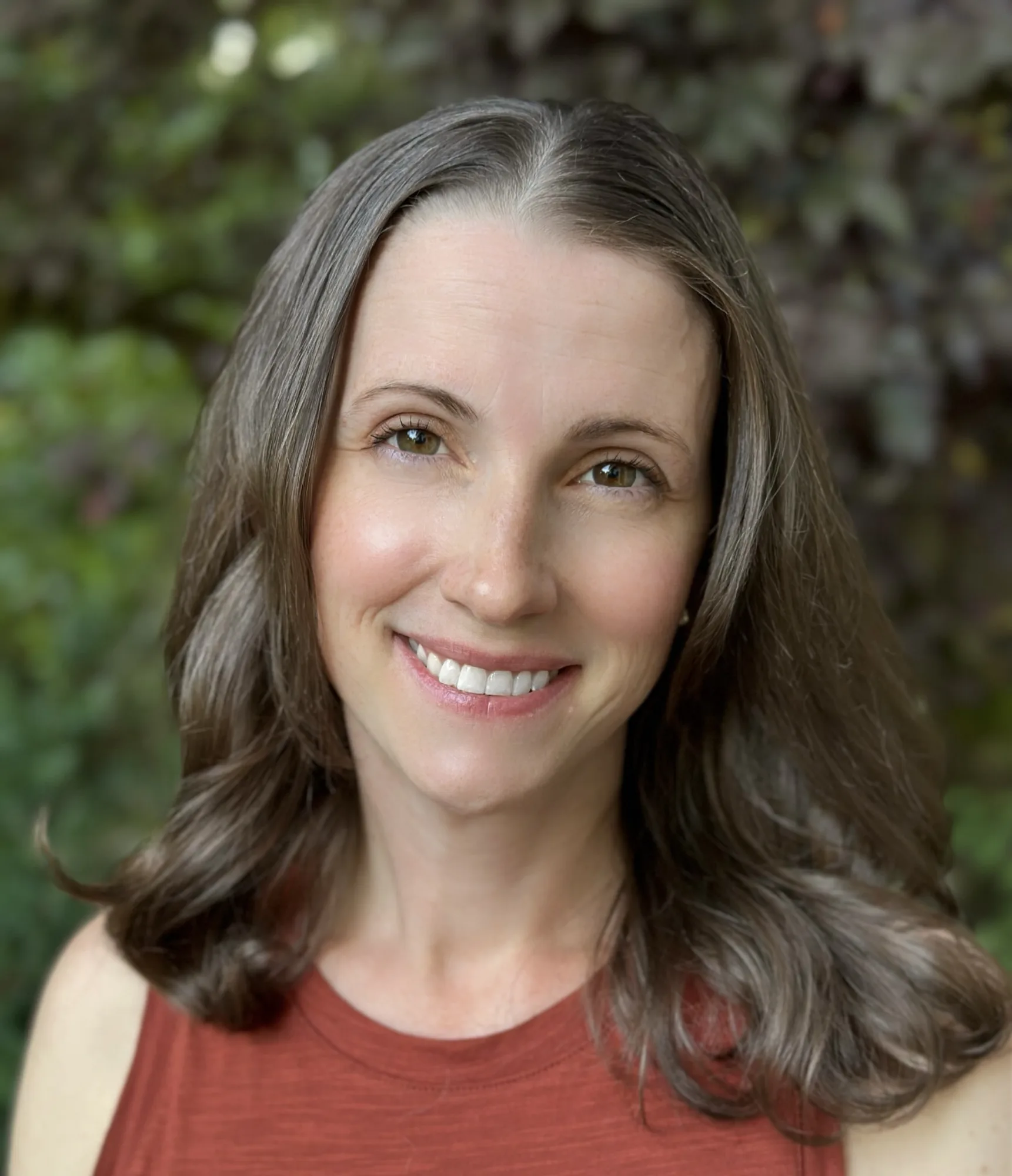 Woman with long brown hair wearing rust-colored top smiling at camera