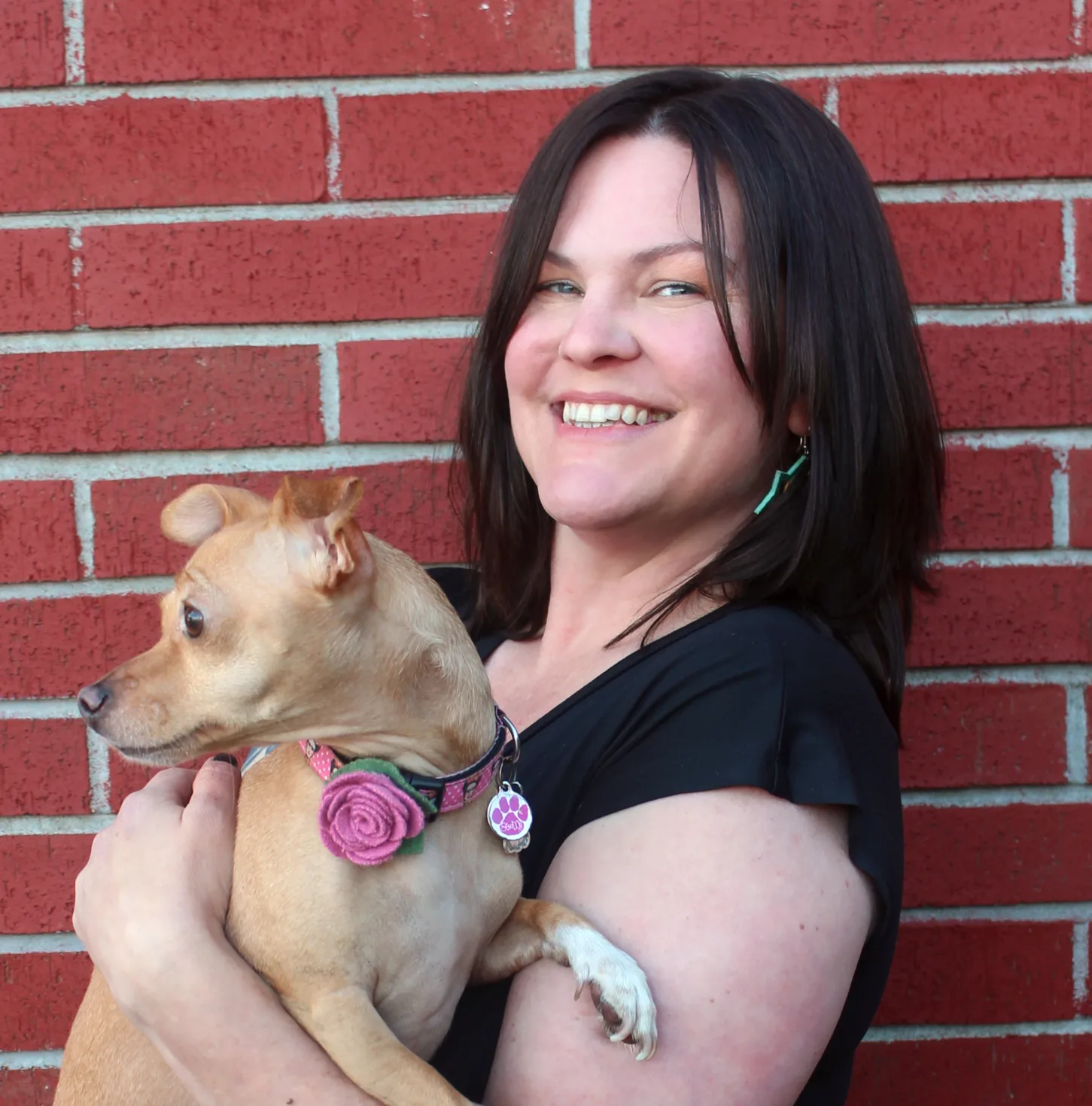Woman with dark hair smiling while holding a small tan dog wearing a pink rose collar