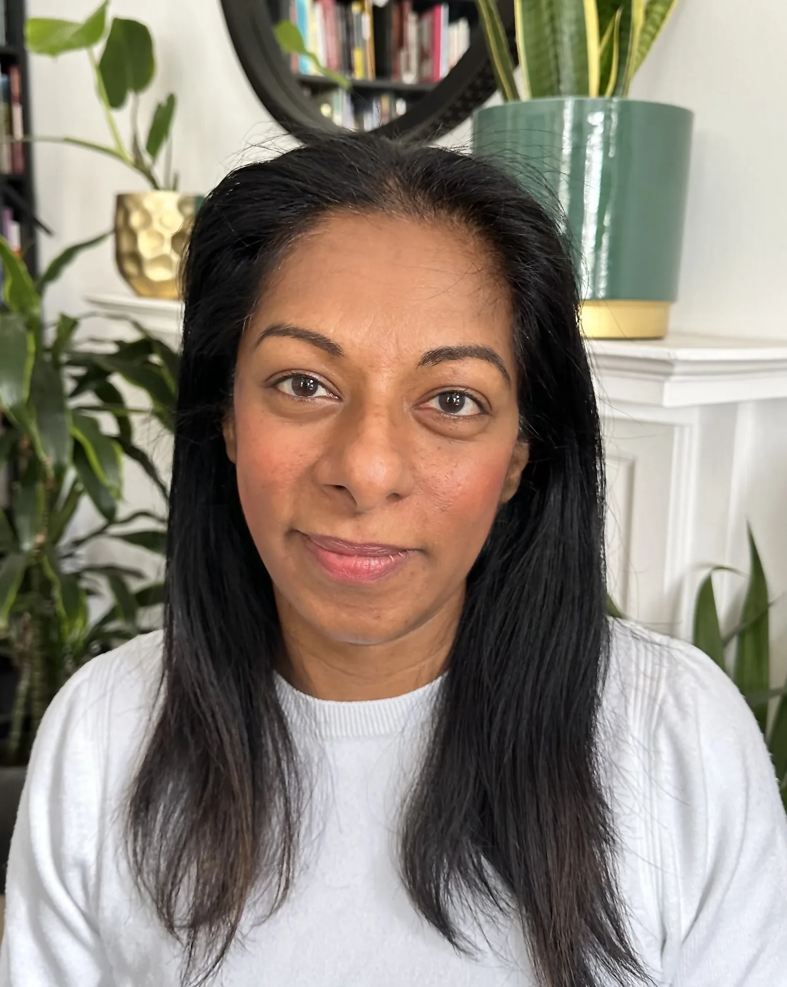 Woman with long dark hair in white shirt seated in home with plants and bookshelf