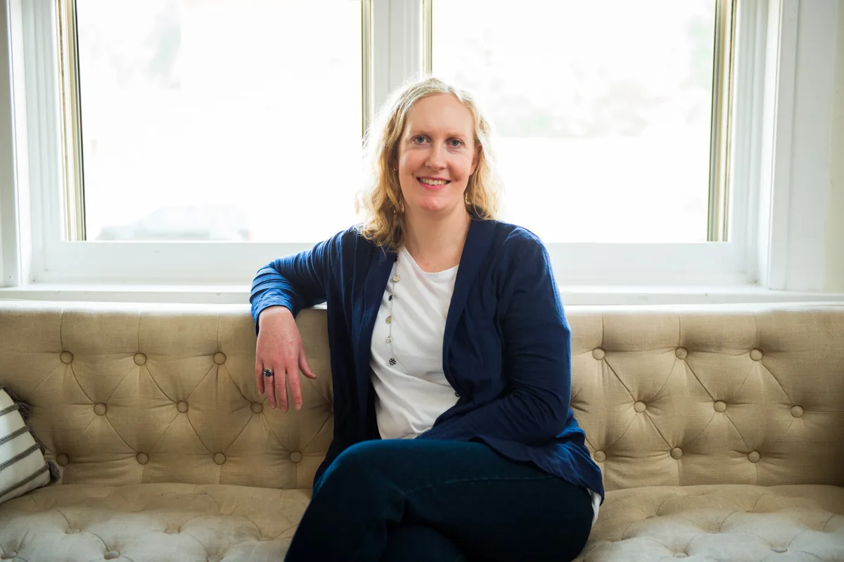 Woman with blonde hair in blue blazer and white top sitting on beige tufted sofa