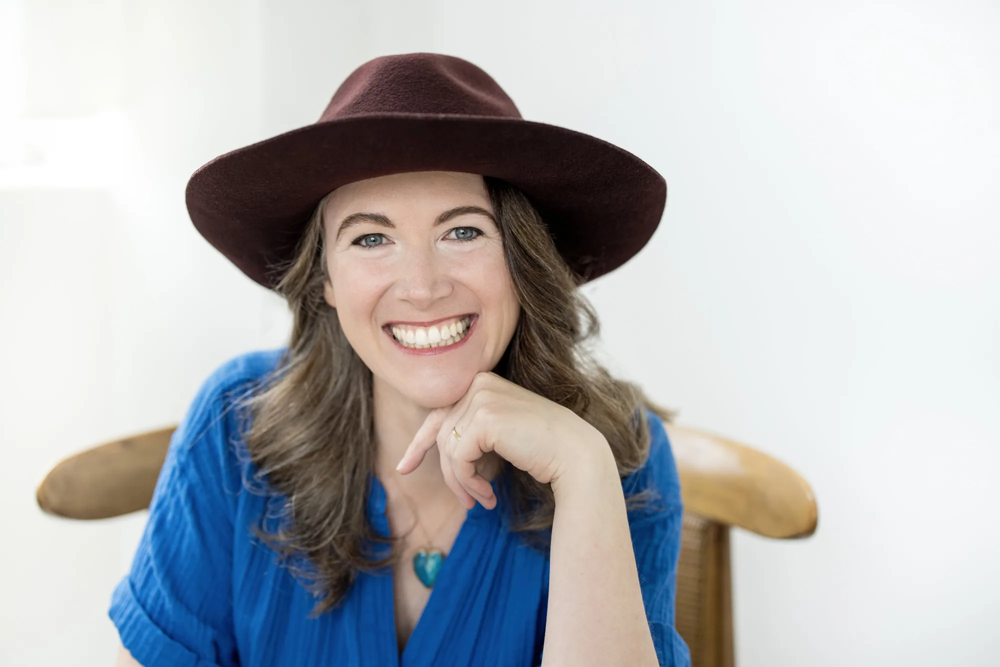 Woman in brown wide-brim hat and blue cardigan smiling with hand under chin