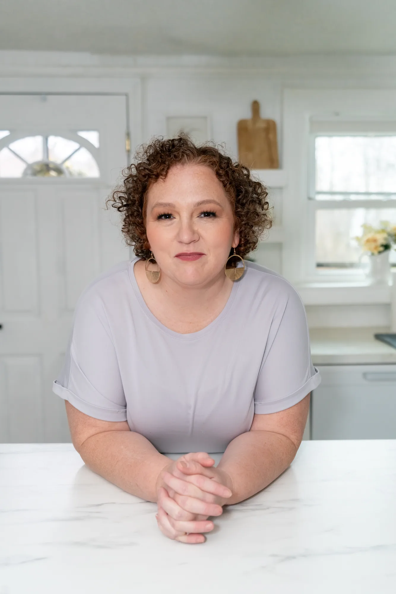 Woman with curly hair wearing gray shirt standing in bright white kitchen