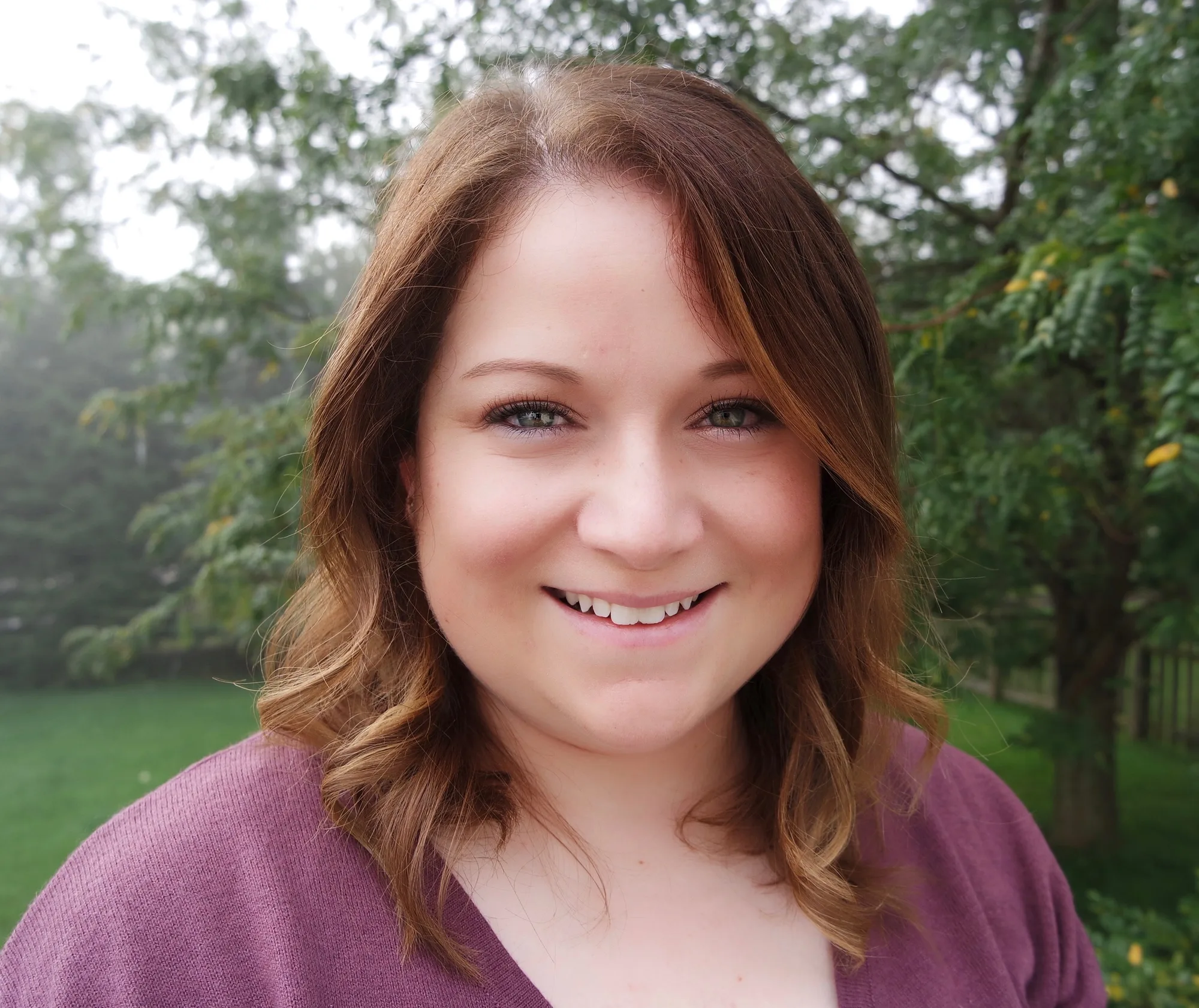 Woman with auburn hair in purple sweater smiling outdoors with trees in background