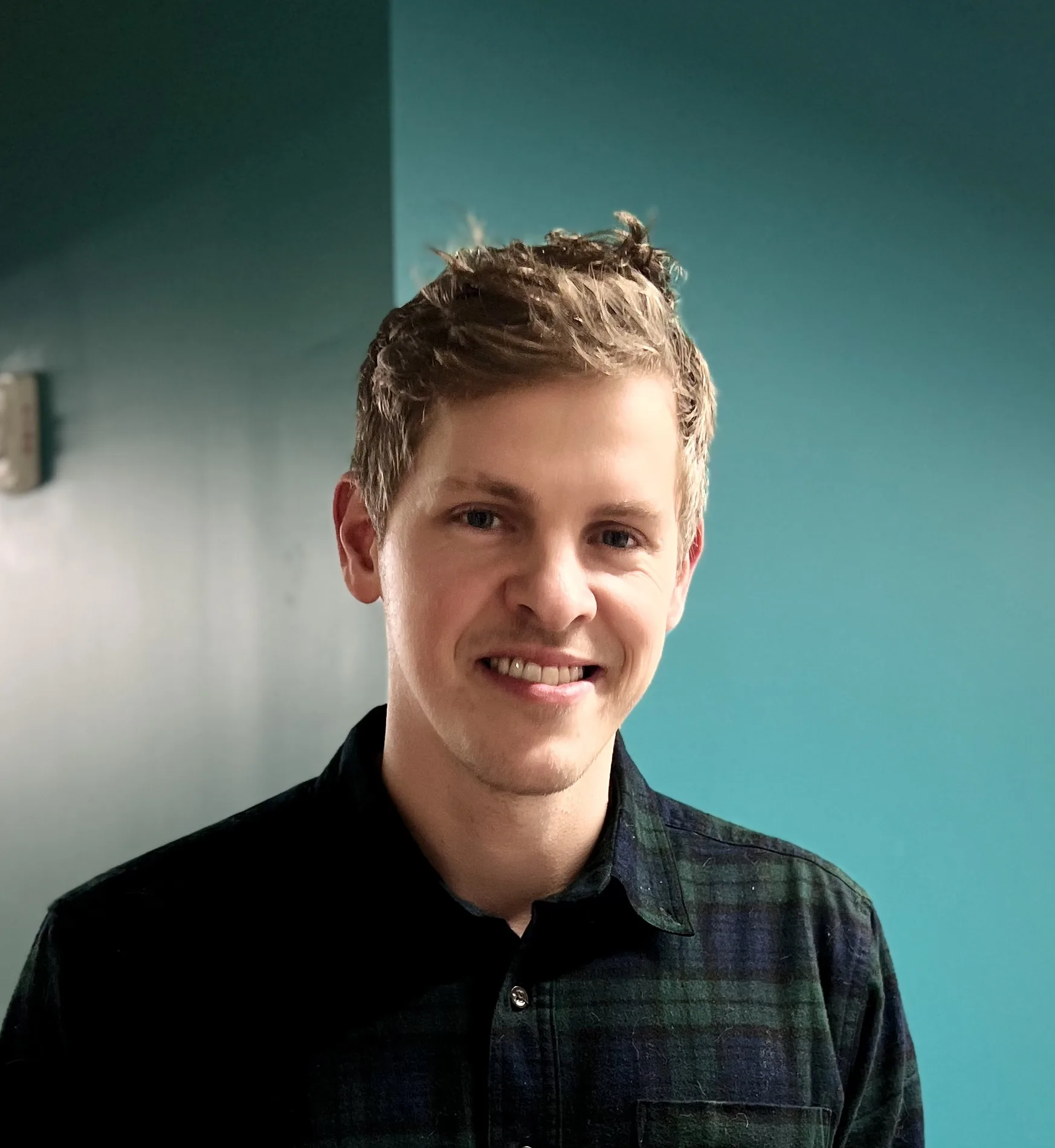 Smiling man with short hair wearing plaid shirt against teal wall